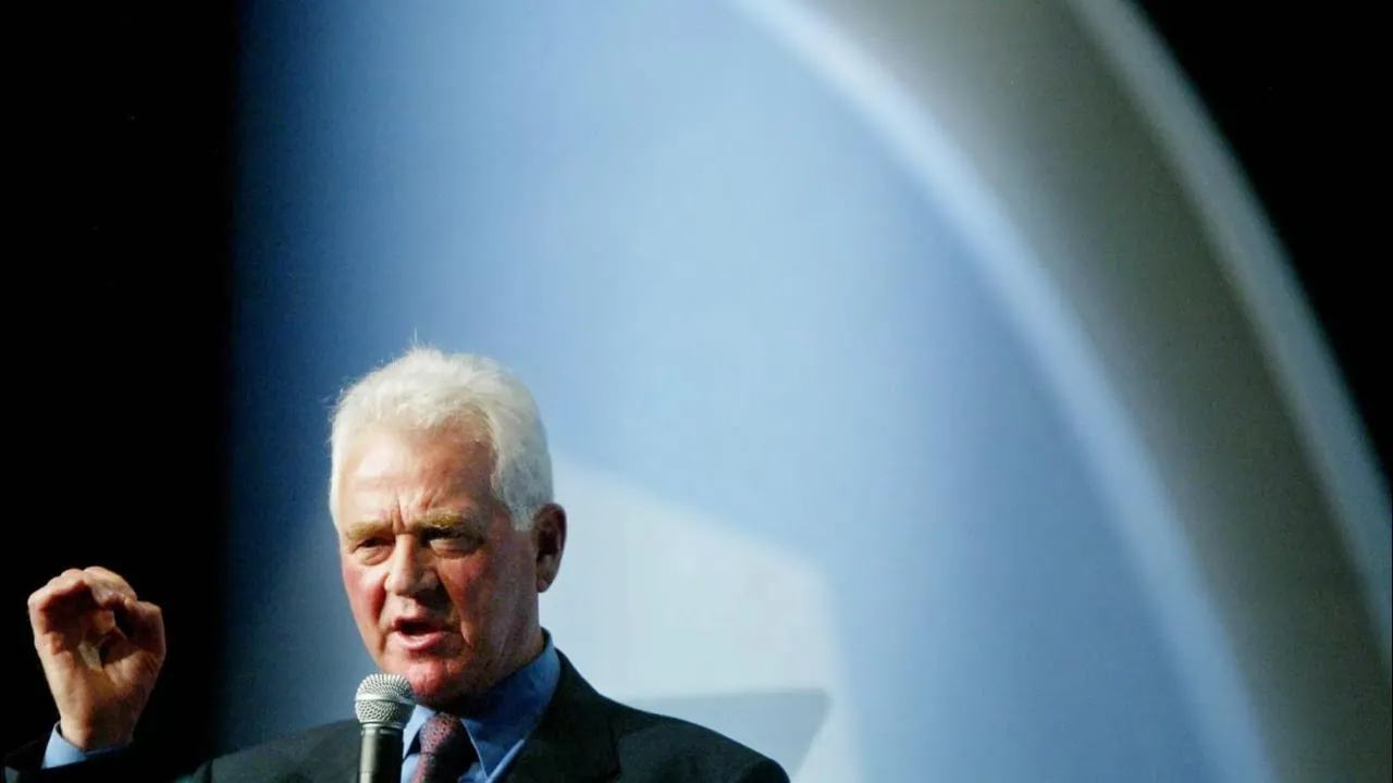Frank Stronach speaking into a microphone on a dark stage