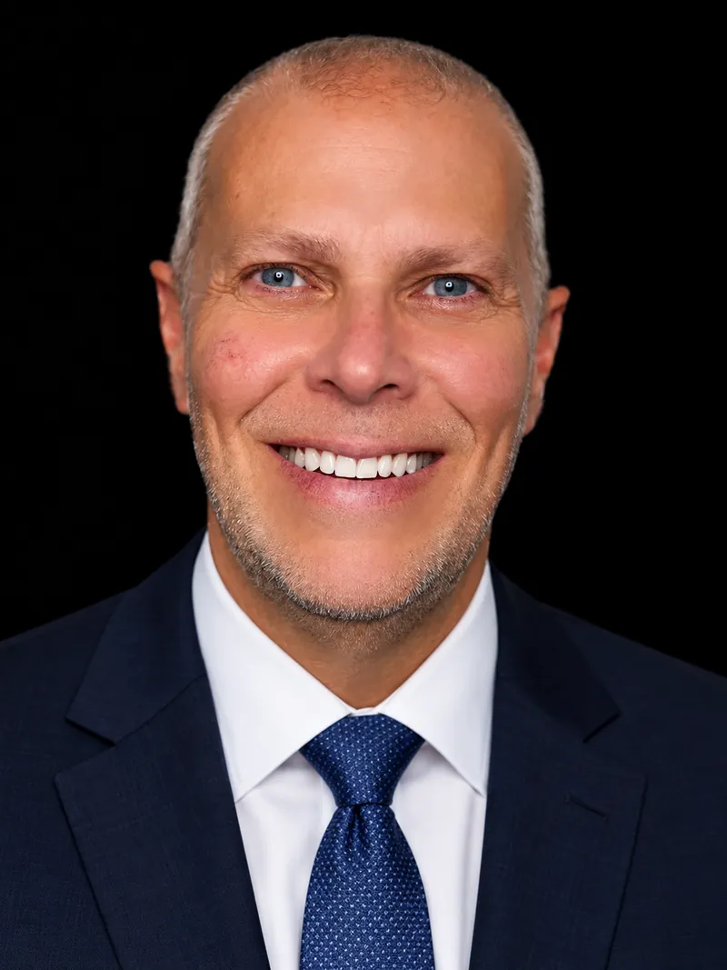 Mike Bayer, head and shoulders, smiling, in a navy suit and tie on a black background